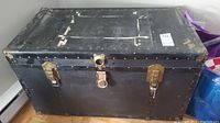 Front view of the black metal steamer trunk showing worn surface and brass lock and clasps
