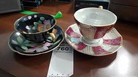 Two vintage teacups and saucers on table showing contrasting designs. One set is iridescent black with floral motif, the other white with red and gold pattern.