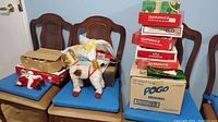 Full view of lot items including several cardboard boxes with ornaments, tinsel, string lights, and plush decorations placed on blue cushions and chairs.