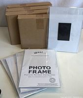 Five brown boxes stacked in the background, five sets of new photo frames in plastic wrap, one frame with black magnetic back visible, on white surface.