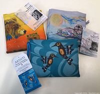 Three folded reusable shopping bags with Indigenous art prints including bears, moonlit landscape with animal, and hummingbirds with tags visible.