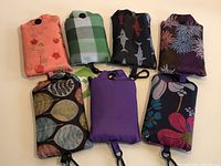 Photo showing seven foldable reusable shopping tote bags in their compact pouches arranged side by side with visible patterns including floral, plaid, sharks, and solid purple.