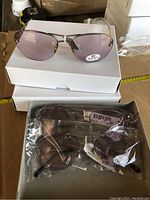 Photo showing three white boxes stacked, each box likely contains three pairs of sunglasses, with one pair on top of the boxes, showing purple tinted lenses and metal frame design.