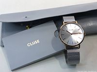 Cluse watch lying next to gray case and instruction manual booklet, showing full dial and mesh band detail.