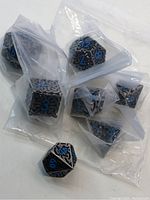 Image showing seven individual polyhedral dice, black and silver metal with blue numbers, each sealed in plastic bags.