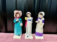 Three Royal Doulton Kate Greenaway figurines side by side on carpeted surface, showing full front view and detail of each figurine's outfit and pose.