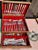 Full view of open wooden chest with red velvet lining displaying various pieces of silverplate flatware including forks, knives, spoons, and a silver candle holder to the side.