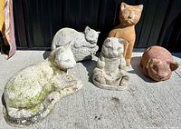 Five garden statues arranged in sunlight on concrete floor: four solid concrete cats showing signs of moss and weathering, one reddish terra cotta pig