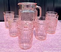 Pitcher and six matching glasses displayed together on a pink surface, showing the pink tint and floral pattern in natural light.