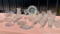Overview front angle showing various types of pressed glassware including goblets, pitchers, plates, and bowls arranged on a pink surface.