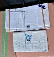 Full set of two folded white cotton linen bundles, one with white-on-white embroidery and one with multi-colored floral embroidery, both tied with ribbons and showing original tags.