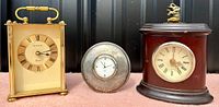 Photo shows three clocks: a Birks quartz carriage clock in brass gold-tone with glass panels, a small round clock with a silver-tone frame, and a brown Holt Renfrew table clock with decorative top