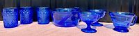 Full set of cobalt blue glassware including tumblers, creamer, dessert dishes, and serving bowl with utensils displayed on a surface.