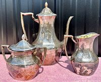 Front view of the full three-piece silver plated coffee set showing the coffee pot, creamer, and sugar bowl with lids, displayed on a pink surface against a black background.