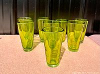 Eight green hand blown glass tumblers arranged on a surface with black metal background, showing their shapes and colors.
