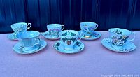 Six vintage tea cups and saucers arranged side by side, showing a variety of floral and crest designs and styles.