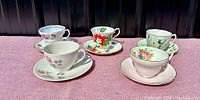 Five vintage tea cups and saucers in various floral designs and colors, arranged on a purple surface.