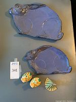 Top view showing large and small purple glazed bunny-shaped platters and three small painted ceramic figures (two bunnies and one butterfly) arranged on a grey surface.