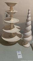 Five stacked ceramic pedestal serving trays alongside a tall stylized ceramic spiral tree sculpture on a gray surface.