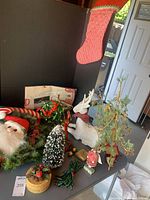 Various Christmas decorations on a table including flocked tree, deer, Santa face, stocking, angel, a basket with pine cones, lights, and ornament hooks