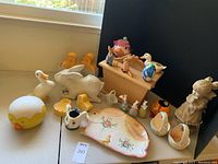 Wide view of Easter-themed porcelain, ceramic, and wooden figurines arranged on a table with visible small yellow chick, white duck, and ceramic plate with floral design.