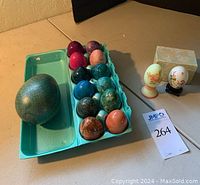 Twelve assorted polished stone eggs arranged in a blue egg carton tray with one large polished stone egg in the tray, next to two painted eggs on stands and a decorative box.