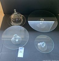 Four clear glass cake plates displayed on a black surface including a covered dessert plate with a dome cover and three pedestal cake plates of various sizes.