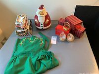 Lot displayed on table including Gumdrop Farmhouse ceramic house, Santa cookie jar, gold-tone baskets in boxes, and green apron.