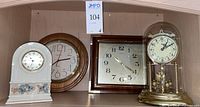 Four clocks displayed on shelf: white ceramic Lenox clock with leaf decoration, round wood-framed clock, square brown clock, and vintage brass and glass dome clock