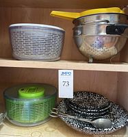 Shelf with two plastic salad spinners, metal colanders (one with cherry design), yellow plastic strainer, and black & white bowls