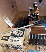 Photo showing Oster electric knife sharpener on top of original box with attached sharpening steel rod, Henckel's wooden knife block holding knives, and black-handled knives behind them.