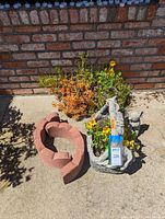 Wide view photo showing the heavy ceramic bunny planter with plants, the metal basket planter with plants, and the red brick scalloped planting border placed on a concrete surface outdoors.