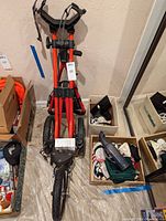 Photo of folded orange and black Sun Mountain Speed Cart Version 1 golf caddy with boxes of golf gloves, a black handheld device, and other golf accessories including a pair of golf shoes.
