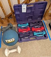 Set of multiple pairs of small hand weights in red, teal, and gray, white coated single dumbbell, and TKO branded medicine ball arranged in a purple case and on carpet