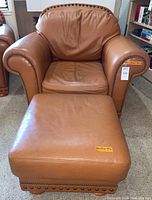 Front view of cinnamon-colored gaucho leather chair and matching ottoman with nailhead trim, both with size tags visible.