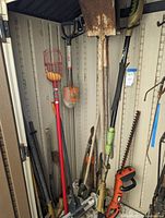 Photo showing long handled shovels, fruit picker, orange electric hedge trimmer, and other garden and hand tools leaning against a shed wall.
