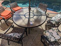 Photo showing full patio set with round glass-top table, 6 chairs with cushions, umbrella in center, by poolside.