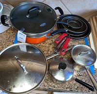 Overview of pots and pans including orange casserole, several fry pans, sauce pans, and lids on granite countertop.