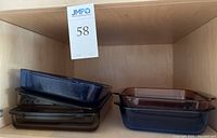 Shelf view of three 13x9 rectangular Pyrex pans stacked, colors include dark smoke, amber, and blue, and two 8x8 square pans in purple and blue stacked to the right