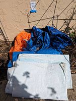 Photo of multiple tarps including a large blue tarp, folded white tarps, and an orange tarp all piled together outdoors.