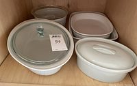 White ceramic bakeware dishes and lids inside a wooden cabinet shelf.