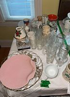 Table with various glassware including beer mugs, stemware, monogrammed glasses, ceramic items like bowls, salt & pepper sets, and serving trays including a pink fan-shaped ceramic tray with gold trim.