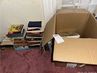 Books stacked on floor and inside a cardboard box, including mixed genres.
