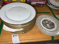 Stack of 12 white plates with a prominent gold rim, partially wrapped with paper towel, showing smooth surfaces and gold detailing.