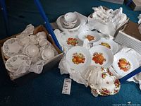 Wide shot showing multiple sets of plates and teacups packed with tissue paper. Includes fruit-themed plates and floral plates.