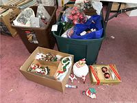 Large plastic bins filled with assorted Christmas ornaments, garland, and decorations, along with a white ceramic Santa cookie jar and some loose items on the floor.