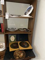 Shelf with multiple collectible plates including two plates in display boxes and several flat plates stacked or displayed on shelves.