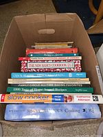 Box of cookbooks stacked with titles clearly visible including 'Slow Cooker Recipe Book' and 'Simply Delicious Cooking'.