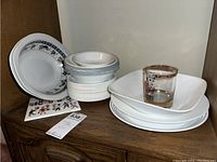 Stack of Corelle white bread plates with floral or blue decorations, stacked bowls, and two rectangular white serving platters with one large serving bowl. Lucy glass with gold rim on top.