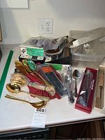 Wide view of assorted flatware and serving utensils laid out on counter, showing loose gold-toned Gotham pieces and boxed International DeepSilver pieces